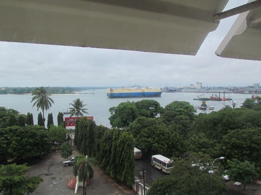 A view of a harbor featuring a large blue cargo ship and various smaller boats, surrounded by lush green trees and palm trees with a cloudy sky above.