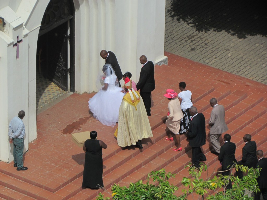 A bride in a white wedding dress walks towards a church entrance, accompanied by a man in a suit. A group of guests in formal attire follows behind, including a man in traditional ceremonial clothing.