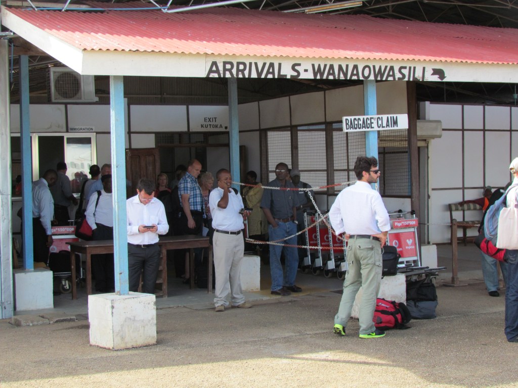 Arrival area at a small airport, featuring a sign for 'Baggage Claim' and several travelers, some on their phones, waiting for their luggage.