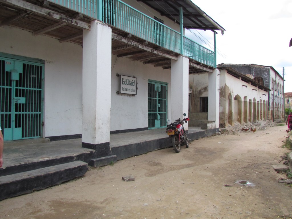 A view of a weathered building with turquoise balcony railings and a sign reading 'EdUkaid' on a dirt road. A parked motorcycle is visible in front, surrounded by abandoned structures.