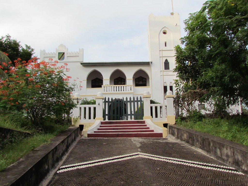 A white building with gothic architectural features, surrounded by greenery and vibrant flowers, leading up to a set of stairs and a wooden gate.