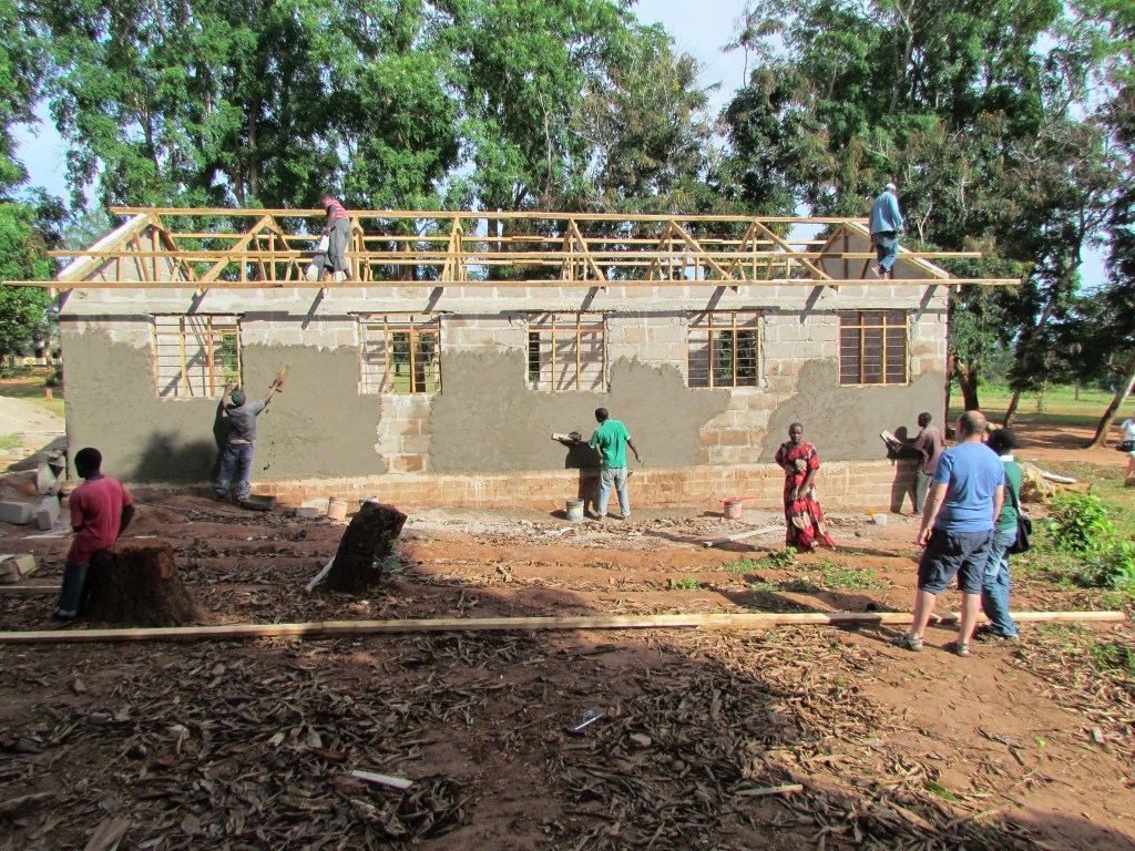Construction workers applying plaster to an incomplete building with a wooden roof structure, surrounded by trees and a construction site.