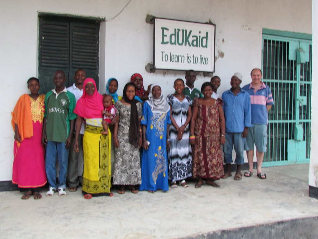 Group photo of diverse individuals in front of a building with an EdUKaid sign, promoting learning and community support.