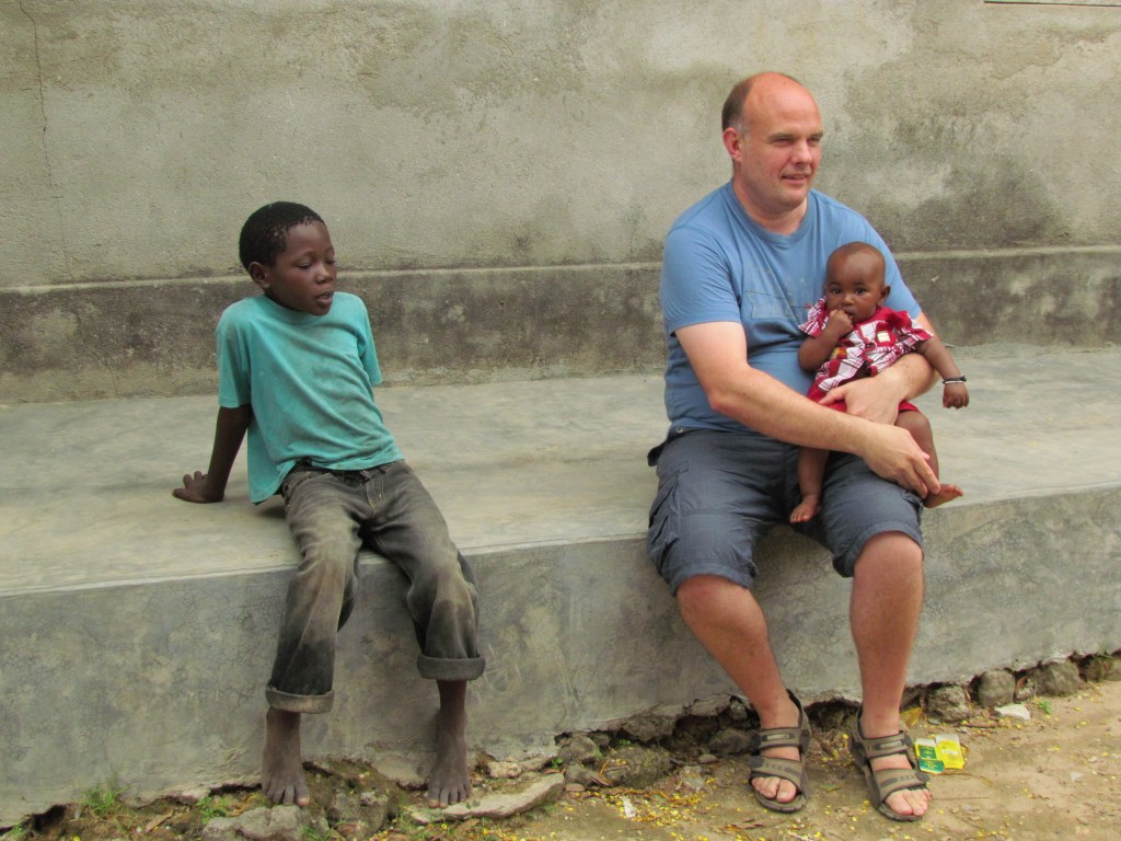 A man sitting on a concrete bench holding a baby in a red outfit, with a young boy in a green shirt seated next to him, against a plain wall.