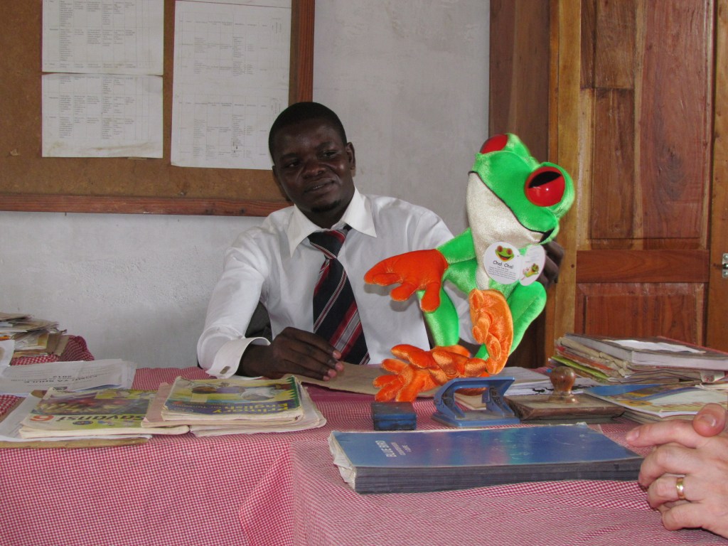 A man in a white shirt and tie sits at a table covered with documents, holding a colorful frog puppet with red eyes and green body.
