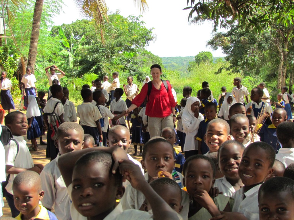 A group of smiling children in school uniforms gathered outdoors, with a woman in a red shirt among them, surrounded by greenery and trees.