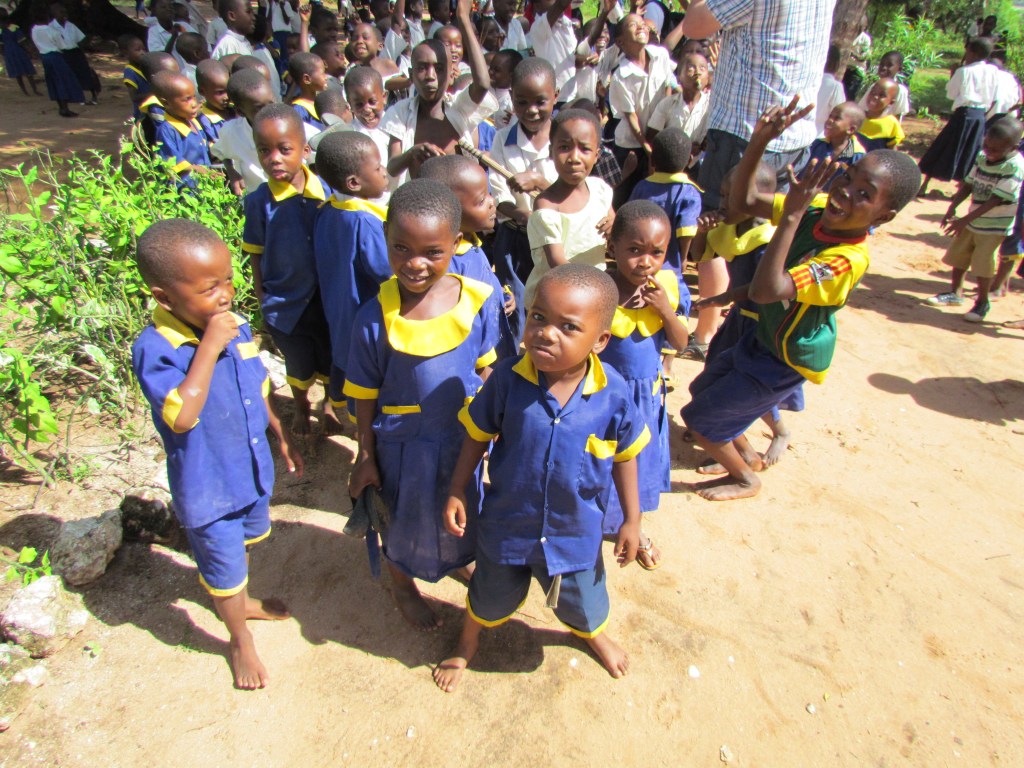 A group of young children in colorful school uniforms smiling and posing for the camera, with a larger crowd of students in the background.