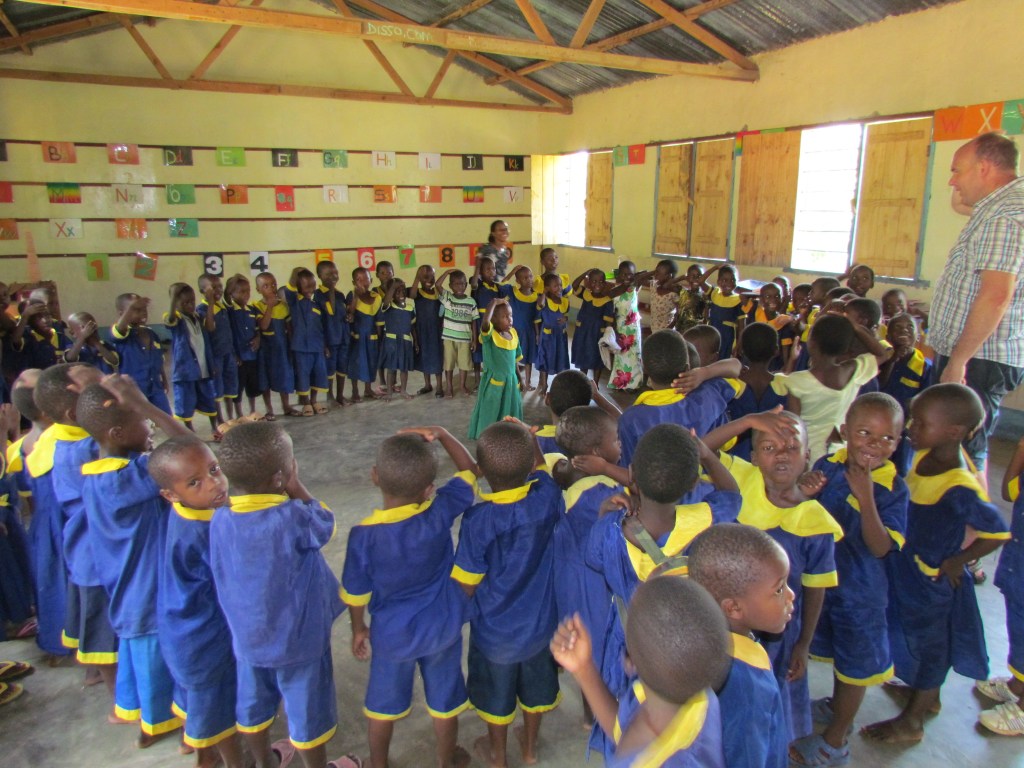 A classroom filled with young children wearing blue and yellow uniforms, engaged in a group activity, while two adults observe. Colorful educational materials are displayed on the walls.