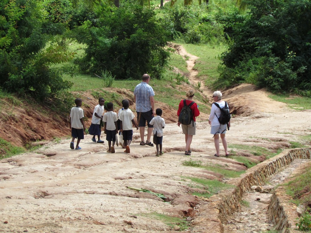 A group of children walking on a dirt path with two adults in a green, rural setting.