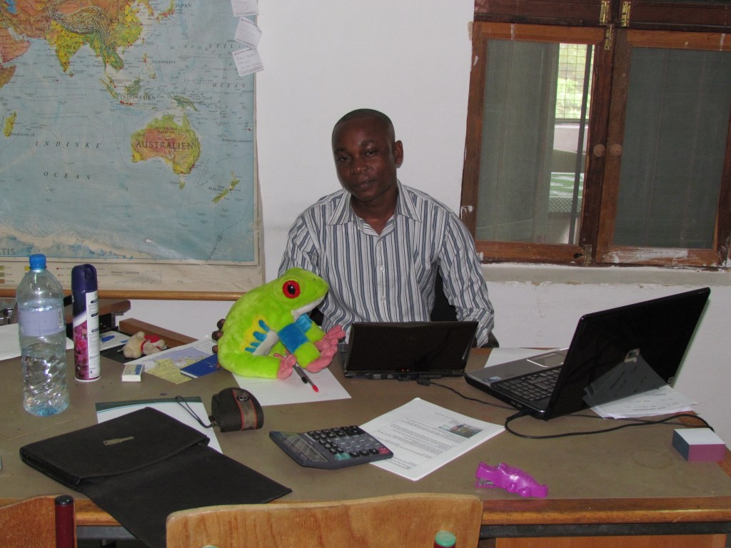 A man sitting at a desk with a large world map in the background. He has a green stuffed frog toy on the table alongside a laptop, a calculator, and various office supplies.