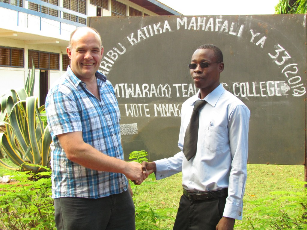 A smiling man in a plaid shirt shakes hands with another man in formal attire in front of a sign for 'Mtiwa B' Teacher's College' surrounded by greenery.
