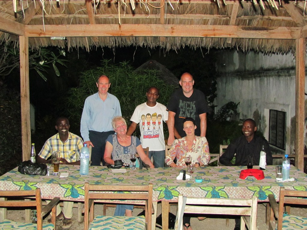 A group of seven people gathered around a table with a colorful tablecloth in an outdoor setting under a thatched roof, with greenery in the background.